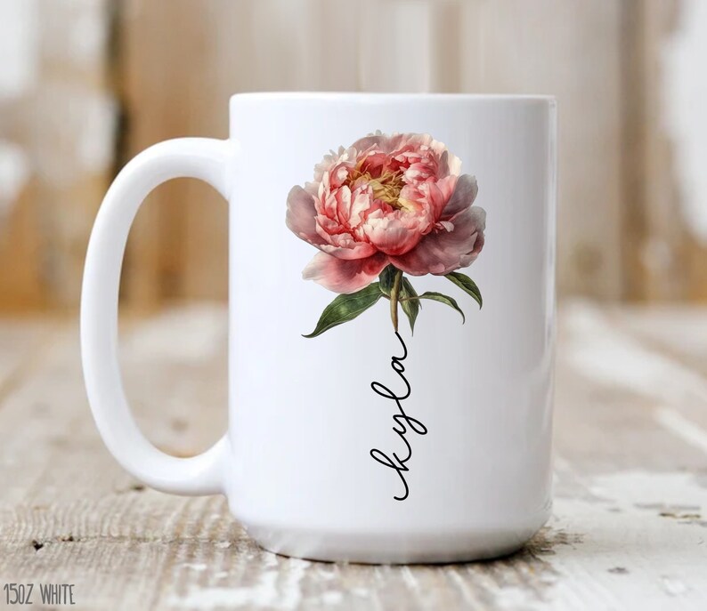 May include: White ceramic mug with a pink peony flower and the name "Kyla" written in black cursive.