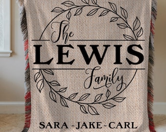 May include: A beige woven throw blanket with black text and a decorative leaf design. The text reads "The LEWIS Family" with the names "SARA - JAKE - CARL" below. The blanket has a fringed edge with red and black accents.