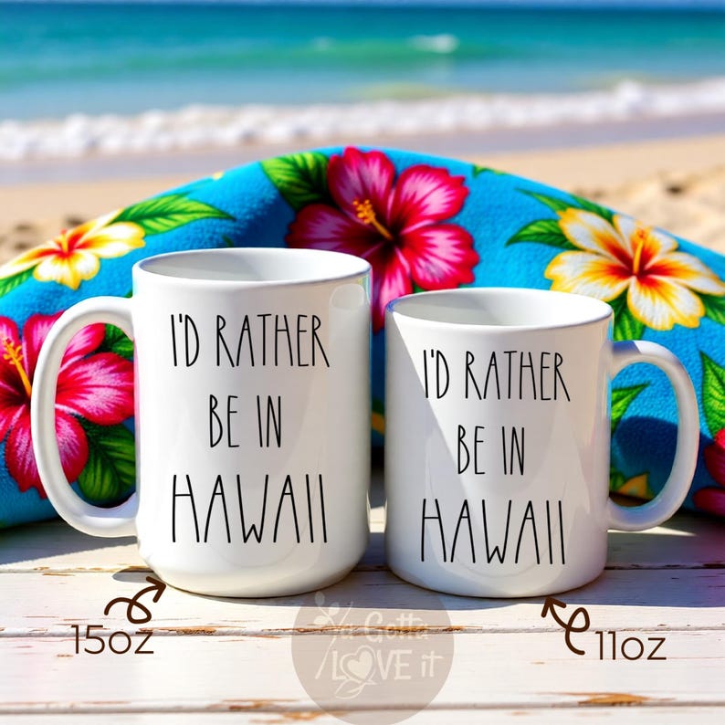 May include: Two white ceramic mugs with the text "I'd Rather Be in Hawaii" in black. One mug is 15oz, the other is 11oz. The mugs are set against a blue floral beach towel with red and yellow flowers.