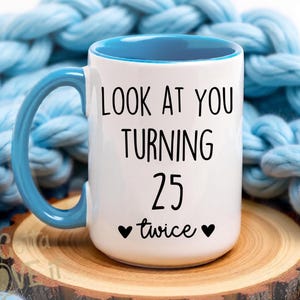May include: A white ceramic mug with a blue handle and interior. The front features black text that reads LOOK AT YOU TURNING 25 twice, flanked by two small black hearts. The mug has a cylindrical body and a curved handle on the left side.