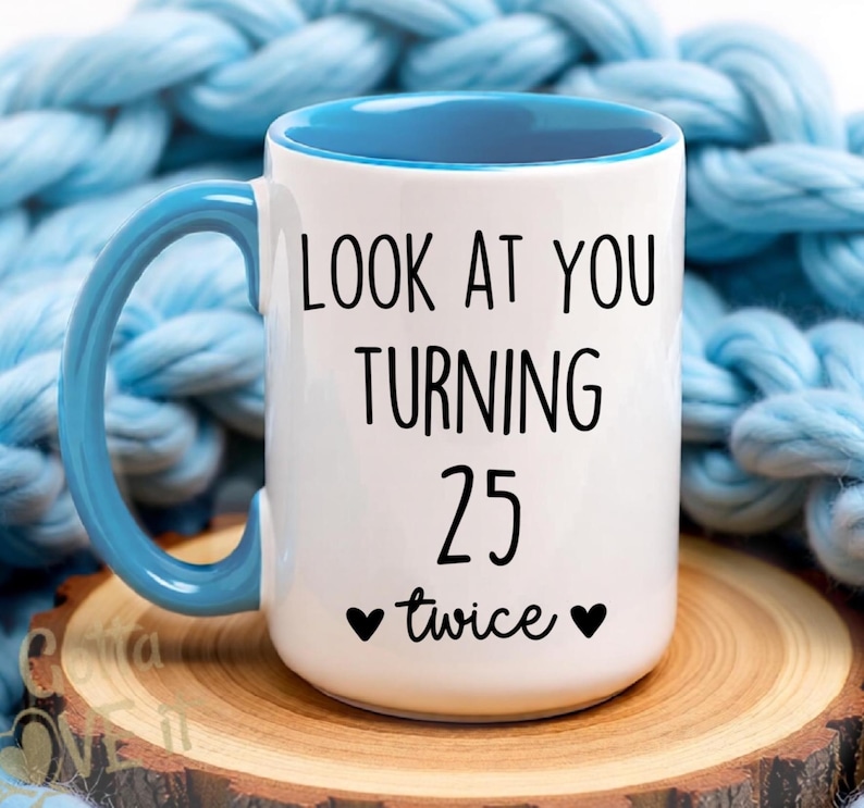May include: A white ceramic mug with a blue handle and interior. The front features black text that reads LOOK AT YOU TURNING 25 twice, flanked by two small black hearts. The mug has a cylindrical body and a curved handle on the left side.