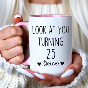 May include: A white ceramic mug with a light pink handle and interior. The front of the mug features black text that reads "LOOK AT YOU TURNING 25 twice" with a small black heart icon on each side of the word twice, printed in a clear font.