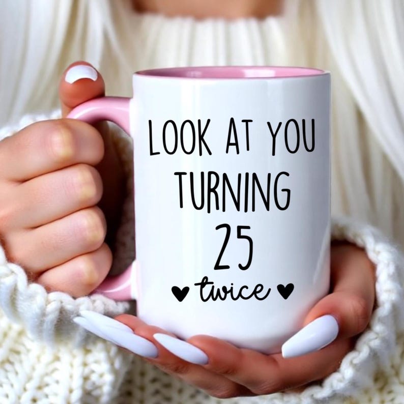 May include: A white ceramic mug with a light pink handle and interior. The front of the mug features black text that reads "LOOK AT YOU TURNING 25 twice" with a small black heart icon on each side of the word twice, printed in a clear font.