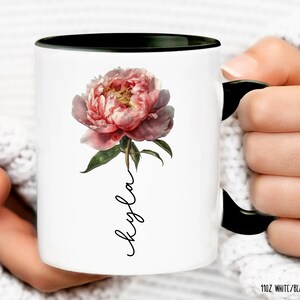 May include: White ceramic mug with a black rim and handle. The mug features a pink peony flower with green leaves and the name "Kayla" written vertically in black script.