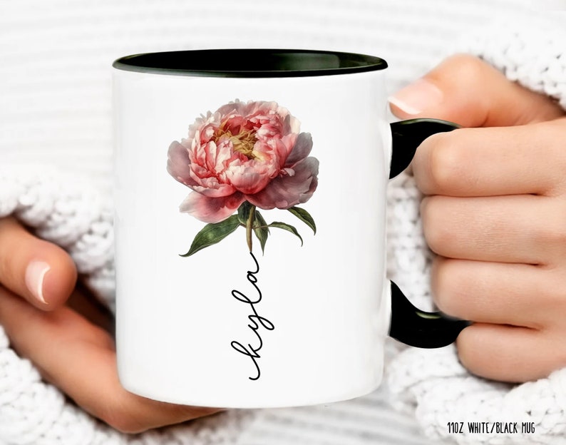 May include: White ceramic mug with a black rim and handle. The mug features a pink peony flower with green leaves and the name "Kayla" written vertically in black script.