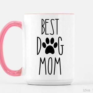 May include: White ceramic mug with a pink handle. The mug has black text that reads "Best Dog Mom" with a paw print in the middle of the word "Dog".