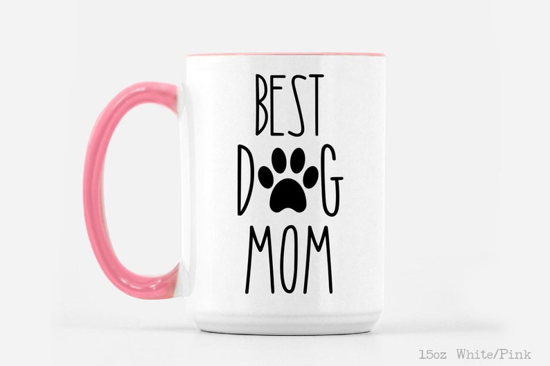 May include: White ceramic mug with a pink handle. The mug has black text that reads "Best Dog Mom" with a paw print in the middle of the word "Dog".