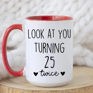 May include: White ceramic mug with a red interior and handle. The mug features the text "LOOK AT YOU TURNING 25 twice" in black, with two black heart symbols. The mug is on a wooden surface.