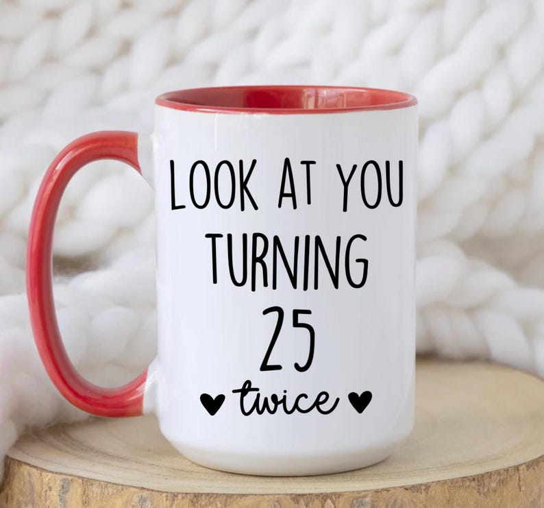 May include: White ceramic mug with a red interior and handle. The mug features the text "LOOK AT YOU TURNING 25 twice" in black, with two black heart symbols. The mug is on a wooden surface.