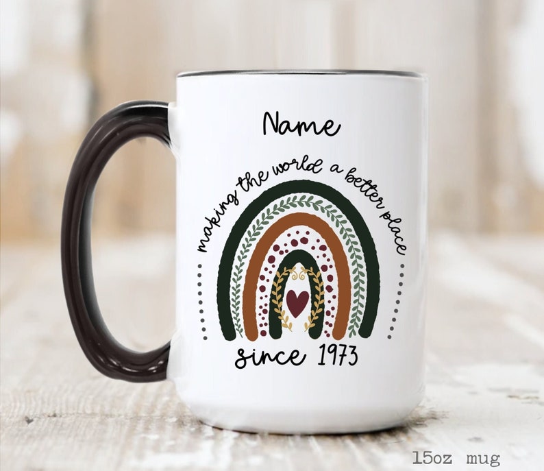 May include: White 15oz mug with a black handle and rim. The mug features a rainbow design with the text "making the world a better place" and "since 1973". The word "Name" is above the rainbow.