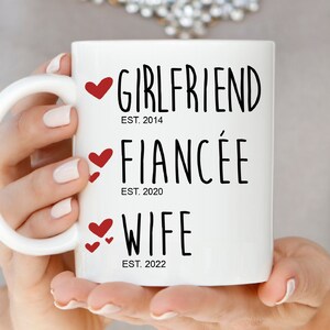以下が含まれることがあります： 白いセラミック製のマグカップに、黒い文字で「Girlfriend Est. 2014、Fianc&eacute;e Est. 2020、Wife Est. 2022」と書いてあります。各行のテキストには赤いハートが付いています。