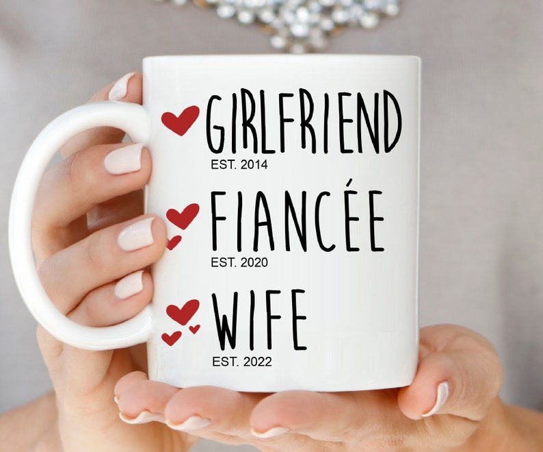 以下が含まれることがあります： 白いセラミック製のマグカップに、黒い文字で「Girlfriend Est. 2014、Fianc&eacute;e Est. 2020、Wife Est. 2022」と書いてあります。各行のテキストには赤いハートが付いています。