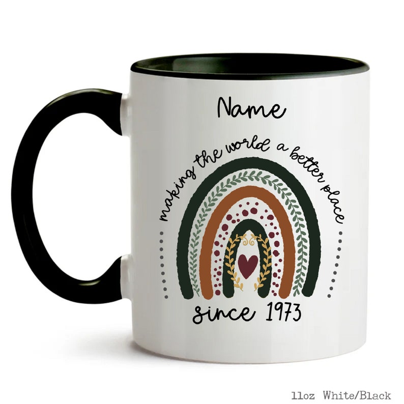 May include: White ceramic mug with a black handle and rim. The mug features a rainbow design with the text "making the world a better place since 1973" and the word "Name" at the top.