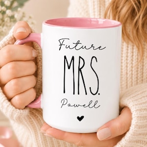 May include: A white ceramic mug with a pink interior and a pink handle. The front of the mug features black text that reads Future MRS. Powell, with a small black heart icon centered below the text. The mug has a smooth, glossy finish.