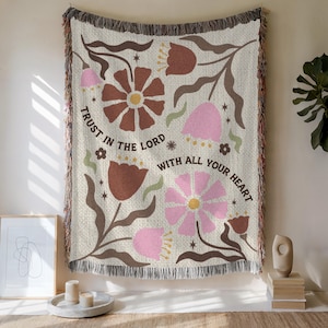 May include: A woven throw blanket featuring a cream background with brown and pink floral illustrations. The text "TRUST IN THE LORD WITH ALL YOUR HEART" is printed in the center. The blanket has a fringed edge with multi-colored threads.