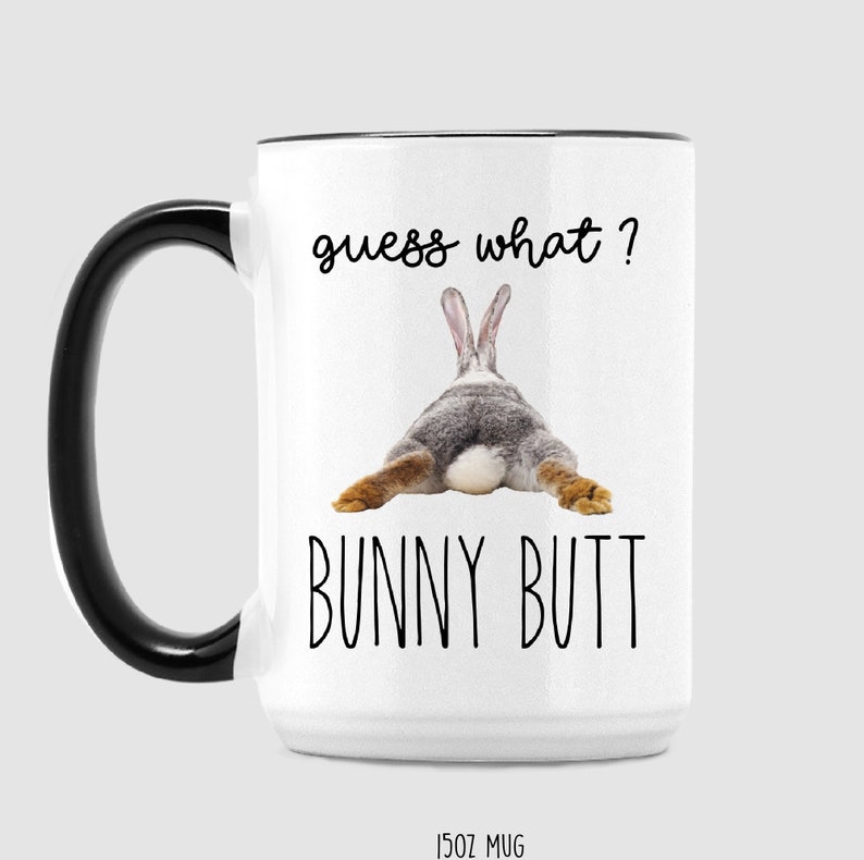 Puede incluir: Taza de cer&aacute;mica blanca con asa y borde negros. La taza presenta el texto "guess what?" encima de una imagen del trasero de un conejo, con las palabras "BUNNY BUTT" debajo. La taza est&aacute; etiquetada como "15OZ MUG".