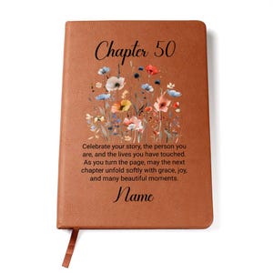 May include: A brown leather journal with the text "Chapter 50" and a floral design. The journal includes the text "Celebrate your story..." and "Name". A brown ribbon bookmark is attached.