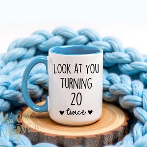 May include: White ceramic mug with a blue interior and handle. The mug has the text "LOOK AT YOU TURNING 20 twice" in black. The mug is on a wooden coaster, with a blue knitted blanket in the background.