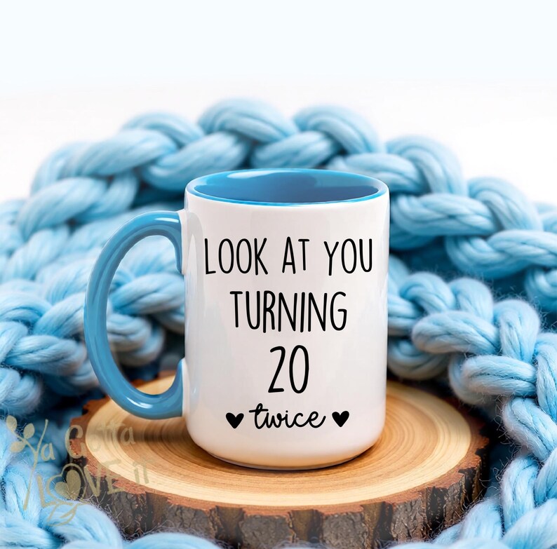 May include: White ceramic mug with a blue interior and handle. The mug has the text "LOOK AT YOU TURNING 20 twice" in black. The mug is on a wooden coaster, with a blue knitted blanket in the background.