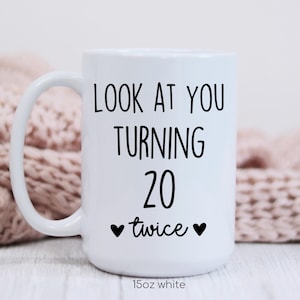 May include: White ceramic mug with a curved handle. The mug features black text that reads "LOOK AT YOU TURNING 20 twice" with two black hearts. The bottom of the mug reads "15oz white".