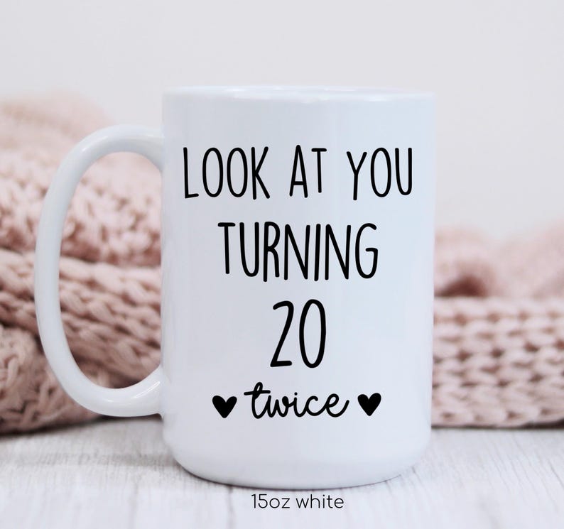 May include: White ceramic mug with a curved handle. The mug features black text that reads "LOOK AT YOU TURNING 20 twice" with two black hearts. The bottom of the mug reads "15oz white".