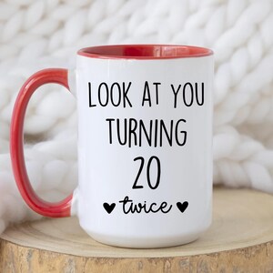 May include: White ceramic mug with a red interior and handle. The mug features the text "LOOK AT YOU TURNING 20 twice" in black, with two black heart symbols. The mug is on a wooden surface.
