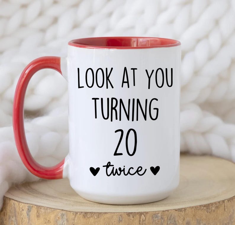 May include: White ceramic mug with a red interior and handle. The mug features the text "LOOK AT YOU TURNING 20 twice" in black, with two black heart symbols. The mug is on a wooden surface.