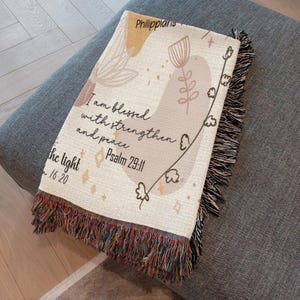 May include: Cream-colored woven throw blanket with a floral design and inspirational text. The blanket features a fringed edge with a mix of colors. The text includes "I am blessed with strength and peace" and "Psalm 29:11".