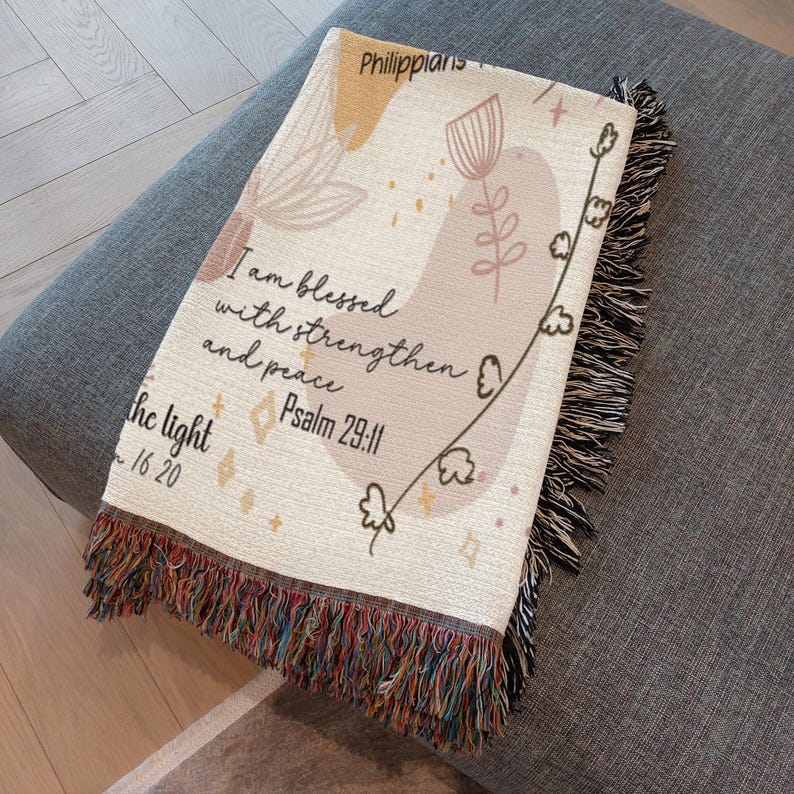 May include: Cream-colored woven throw blanket with a floral design and inspirational text. The blanket features a fringed edge with a mix of colors. The text includes "I am blessed with strength and peace" and "Psalm 29:11".