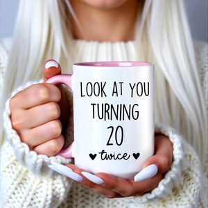 May include: White ceramic mug with a pink interior and handle. The mug features the text "LOOK AT YOU TURNING 20 twice" in black, with two black hearts. The mug is held by a person wearing a white sweater.