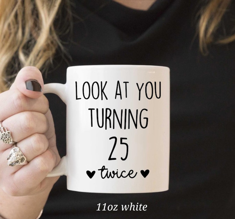 May include: White ceramic mug with the text "LOOK AT YOU TURNING 25 twice" in black. The mug has a black heart on each side of the word "twice". The mug is 11oz.