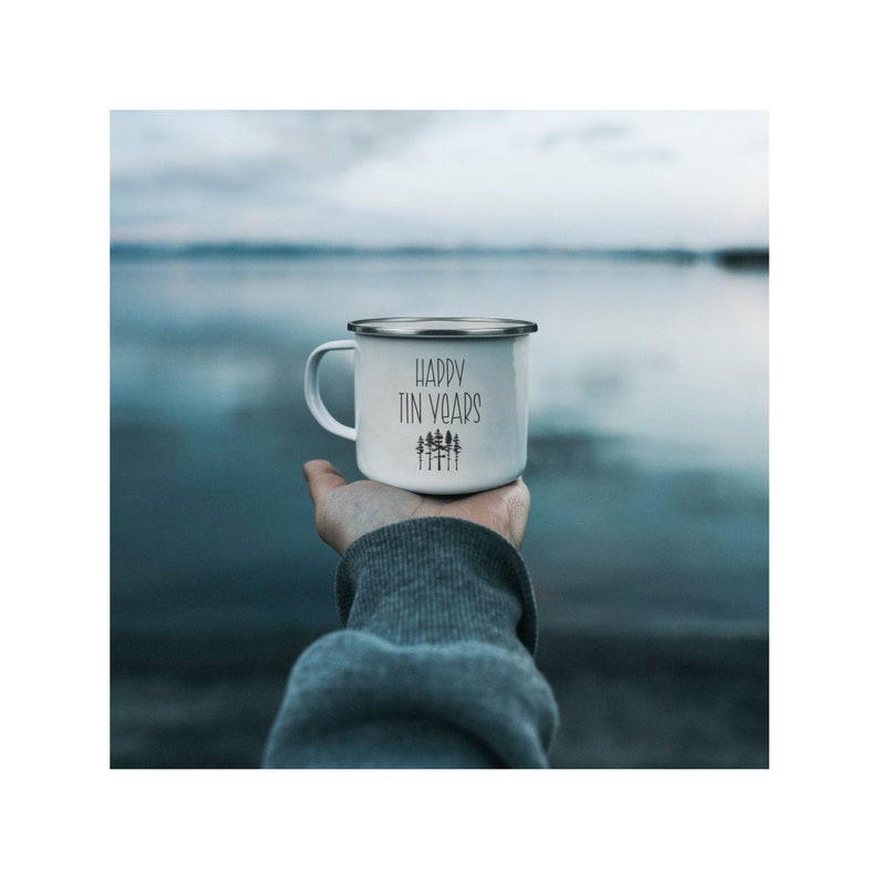 May include: White enamel mug with a black inscription that reads "Happy Tin Years" and a small graphic of a forest. The mug is being held by a hand against a blue and white background.