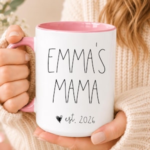 May include: A white ceramic mug with a pink interior and handle. The front features black text that reads EMMA'S MAMA with a small heart icon and the text est. 2026 below it. The mug has a cylindrical shape and a smooth, glossy finish.