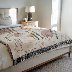 May include: A beige woven throw blanket with floral and botanical designs in muted tones of peach, tan, and brown. The blanket features inspirational text and a black fringe border. The blanket is draped over a bed.
