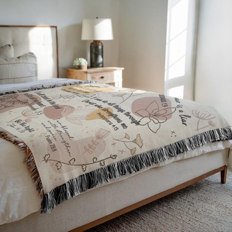 May include: A beige woven throw blanket with floral and botanical designs in muted tones of peach, tan, and brown. The blanket features inspirational text and a black fringe border. The blanket is draped over a bed.