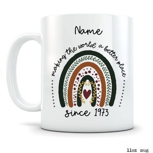 May include: White ceramic mug with a rainbow design in shades of green, brown, and red, with the text "making the world a better place since 1973" and the name "Name". The mug is 11oz.