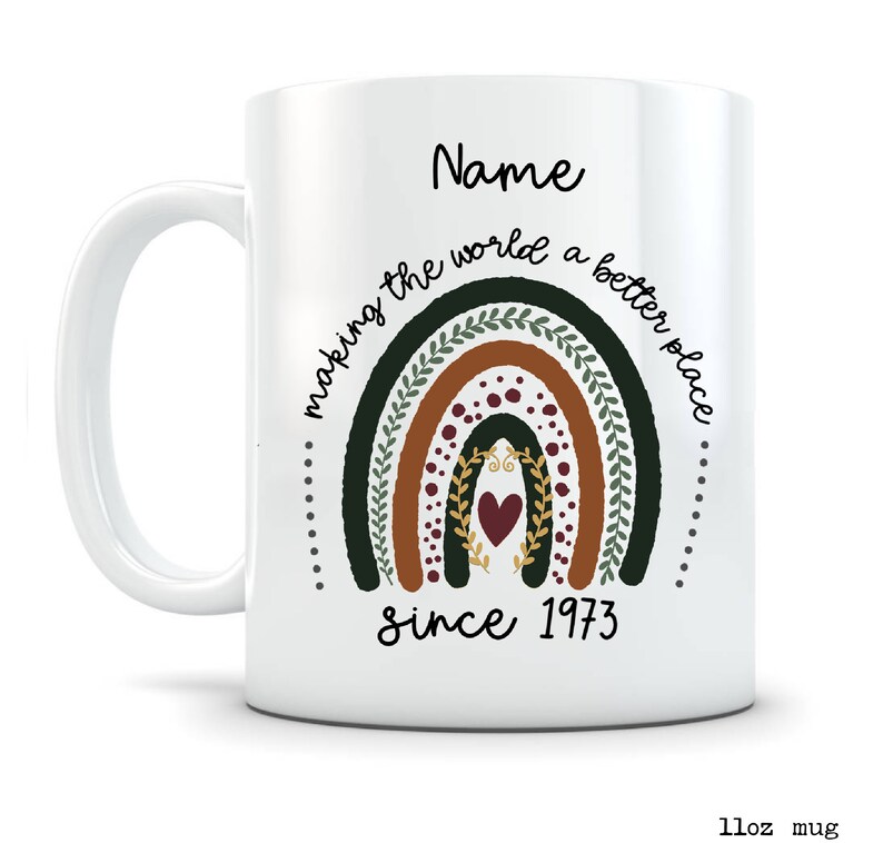 May include: White ceramic mug with a rainbow design in shades of green, brown, and red, with the text "making the world a better place since 1973" and the name "Name". The mug is 11oz.