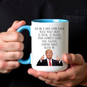 Puede incluir: Taza de cer&aacute;mica blanca con interior y asa azul claro. La taza presenta el texto "YOU ARE A GREAT GUITAR PLAYER. REALLY, REALLY GREAT!" y una imagen de un hombre haciendo una se&ntilde;al de pulgar.