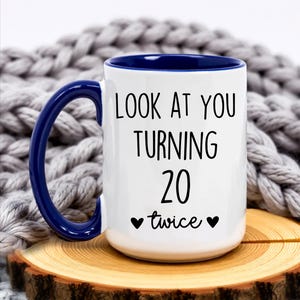 May include: White ceramic mug with a blue handle and interior. The mug features the text "LOOK AT YOU TURNING 20 twice" in black, with two black heart symbols. The mug is on a wooden slice.
