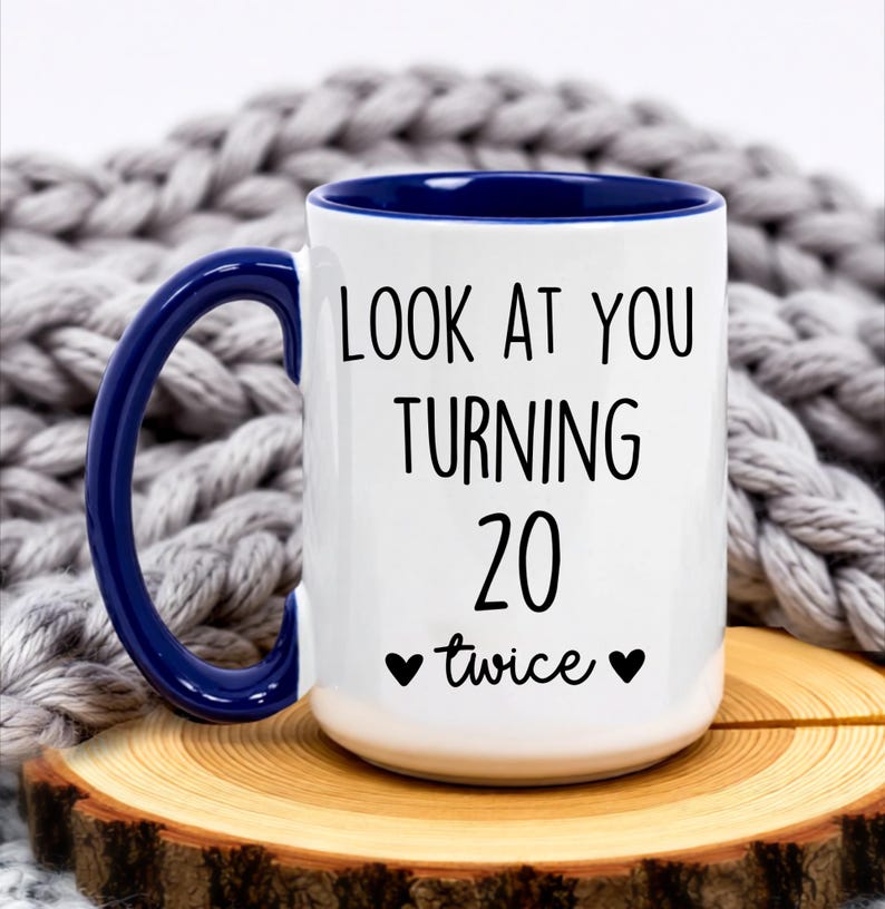 May include: White ceramic mug with a blue handle and interior. The mug features the text "LOOK AT YOU TURNING 20 twice" in black, with two black heart symbols. The mug is on a wooden slice.
