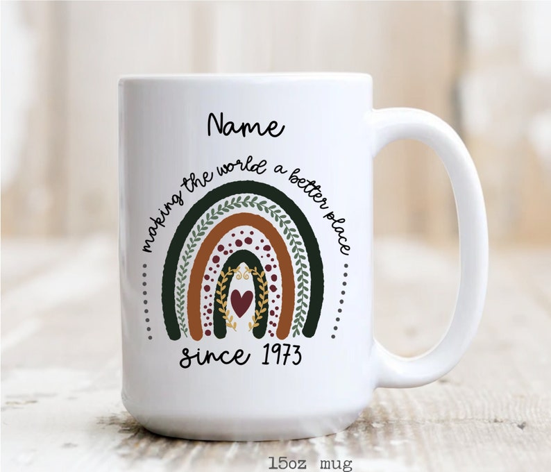 May include: White ceramic mug with a rainbow design in shades of green, brown, and burgundy, with the text "making the world a better place" and "since 1973". The mug is 15oz.