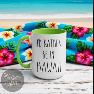 May include: White ceramic mug with the text "I'D RATHER BE IN HAWAII" in black. The mug has a green handle and interior. It is set against a blue floral towel with pink and yellow flowers, with a beach and ocean background.