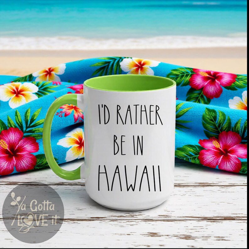 May include: White ceramic mug with the text "I'D RATHER BE IN HAWAII" in black. The mug has a green handle and interior. It is set against a blue floral towel with pink and yellow flowers, with a beach and ocean background.