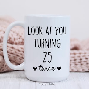 May include: White ceramic mug with a curved handle. The mug features black text that reads "LOOK AT YOU TURNING 25 twice" with two black hearts. The bottom of the mug reads "15oz white".