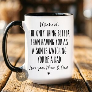 May include: A white ceramic mug with a black handle and interior rim. The mug features black printed text that reads: Michael, THE ONLY THING BETTER THAN HAVING YOU AS A SON IS WATCHING YOU BE A DAD, Love you, Mom & Dad, with a small heart icon below.