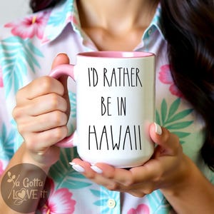 May include: White ceramic mug with a pink handle and interior. The mug features the text "I'D RATHER BE IN HAWAII" in black lettering. The mug is held by a person wearing a floral shirt.