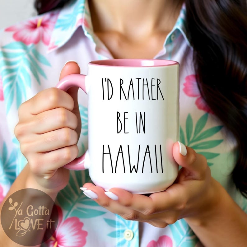 May include: White ceramic mug with a pink handle and interior. The mug features the text "I'D RATHER BE IN HAWAII" in black lettering. The mug is held by a person wearing a floral shirt.