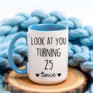 May include: White ceramic mug with a blue handle and interior. The mug features the text "LOOK AT YOU TURNING 25 twice" in black. The mug is on a wooden coaster, with a blue knitted blanket in the background.