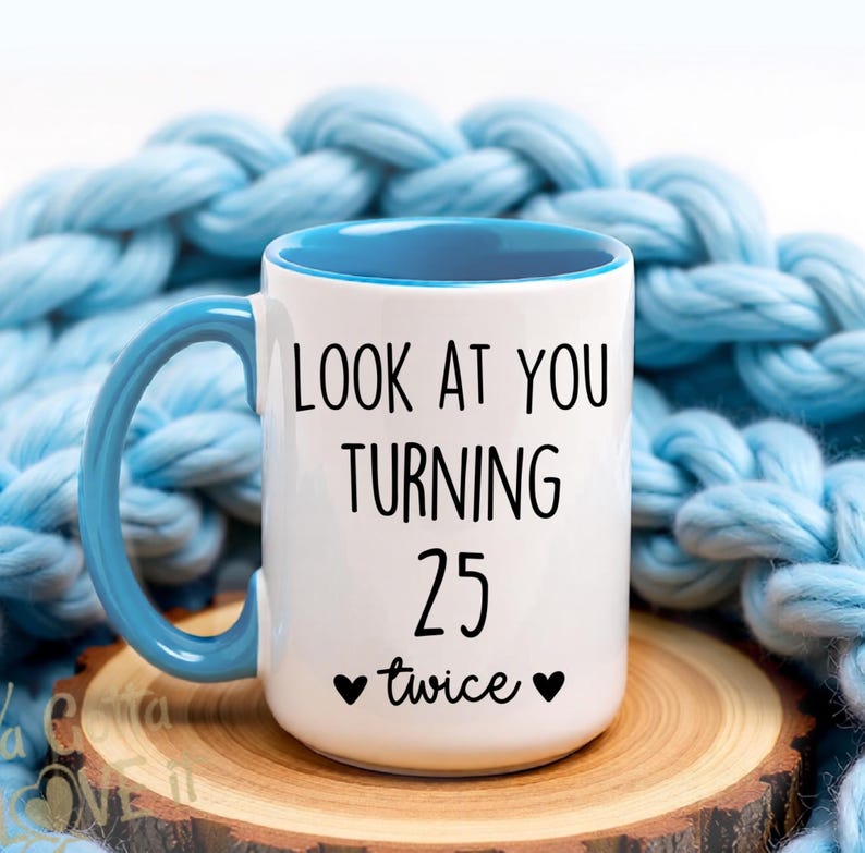 May include: White ceramic mug with a blue handle and interior. The mug features the text "LOOK AT YOU TURNING 25 twice" in black. The mug is on a wooden coaster, with a blue knitted blanket in the background.