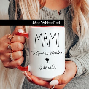 May include: White ceramic mug with a red rim. The mug has black text that reads "MAMI Te Quiero Mucho" with a small heart and the name "Gabriela" underneath.